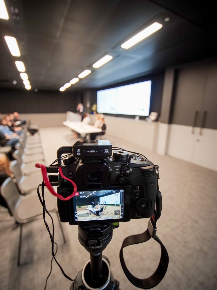 Camera on a tripod recording a boardroom panel session, attendees seated along the table.