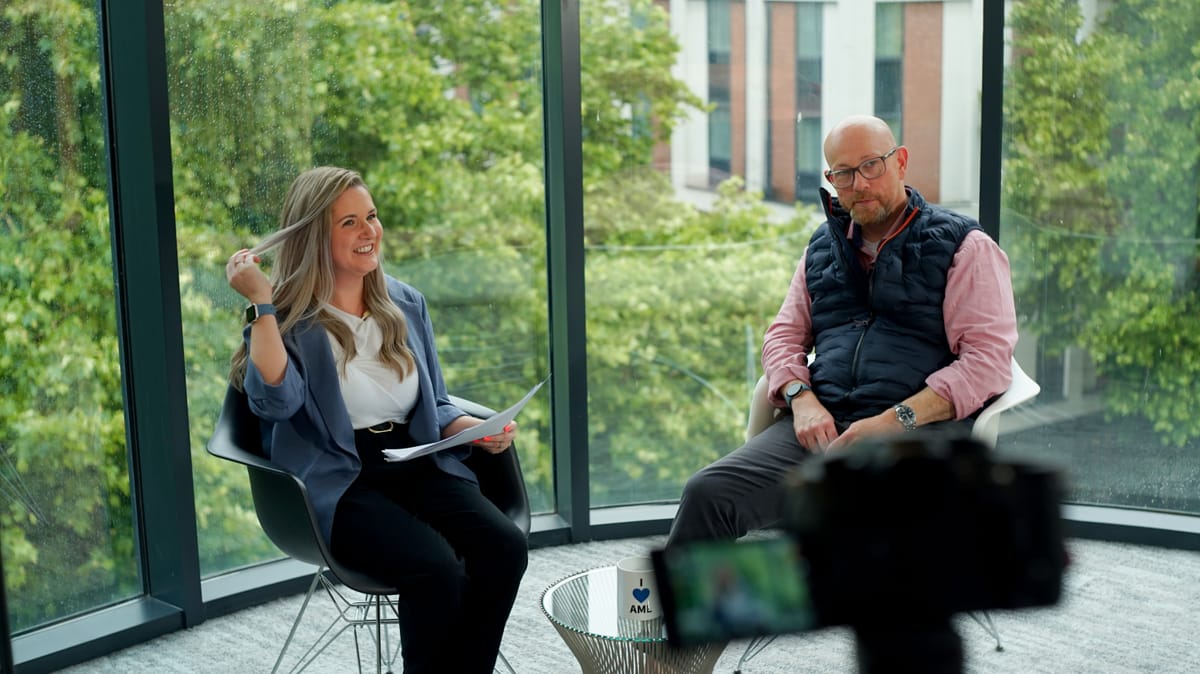 An interview in progress in a glass-walled room, camera framed in the foreground.